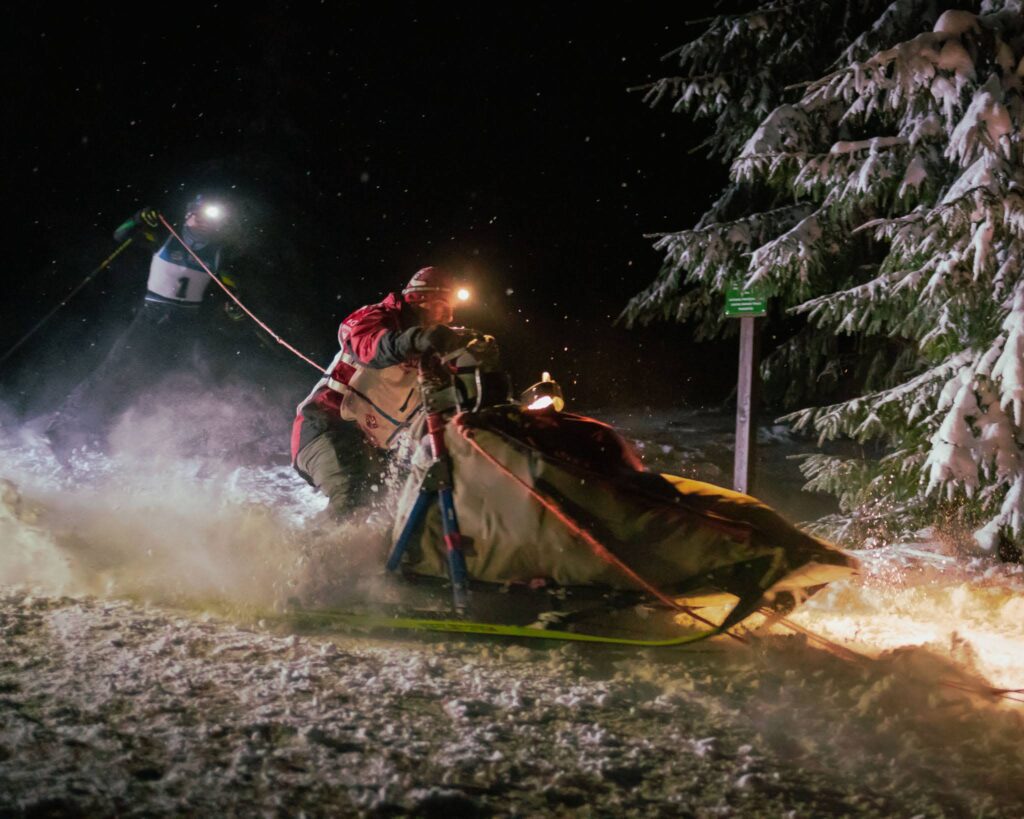 musher sliding on his sled with a skier behind him during the night race