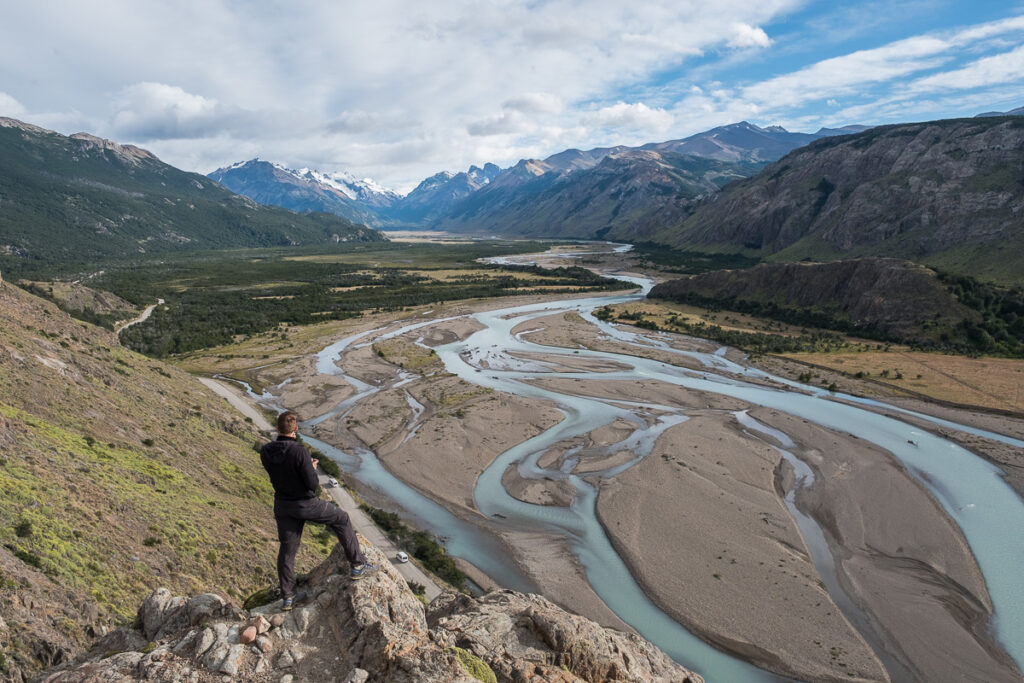 meandering river in the wide valley with snowy peaks in the background. A guy standing on the edge of the cliff falling into the river